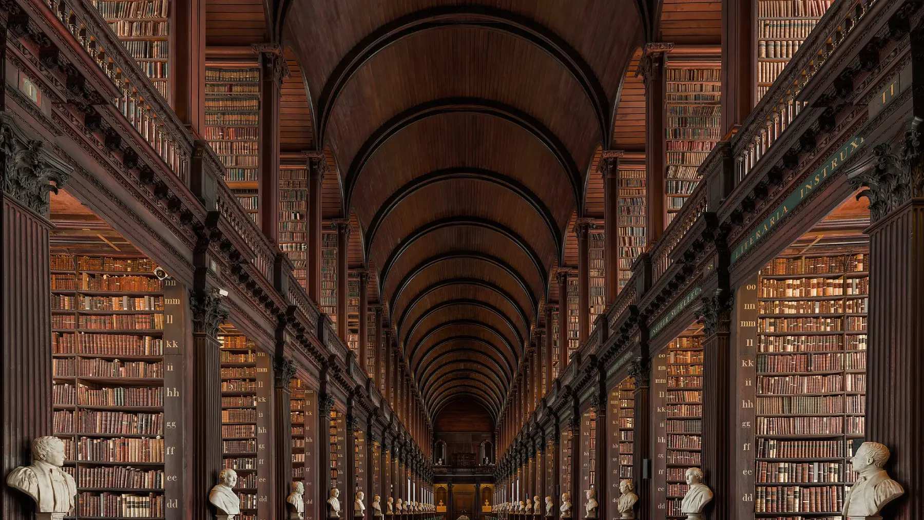 Interior of a grand historic library with high vaulted wooden ceiling, rows of tall bookshelves, and marble busts lining the hall.