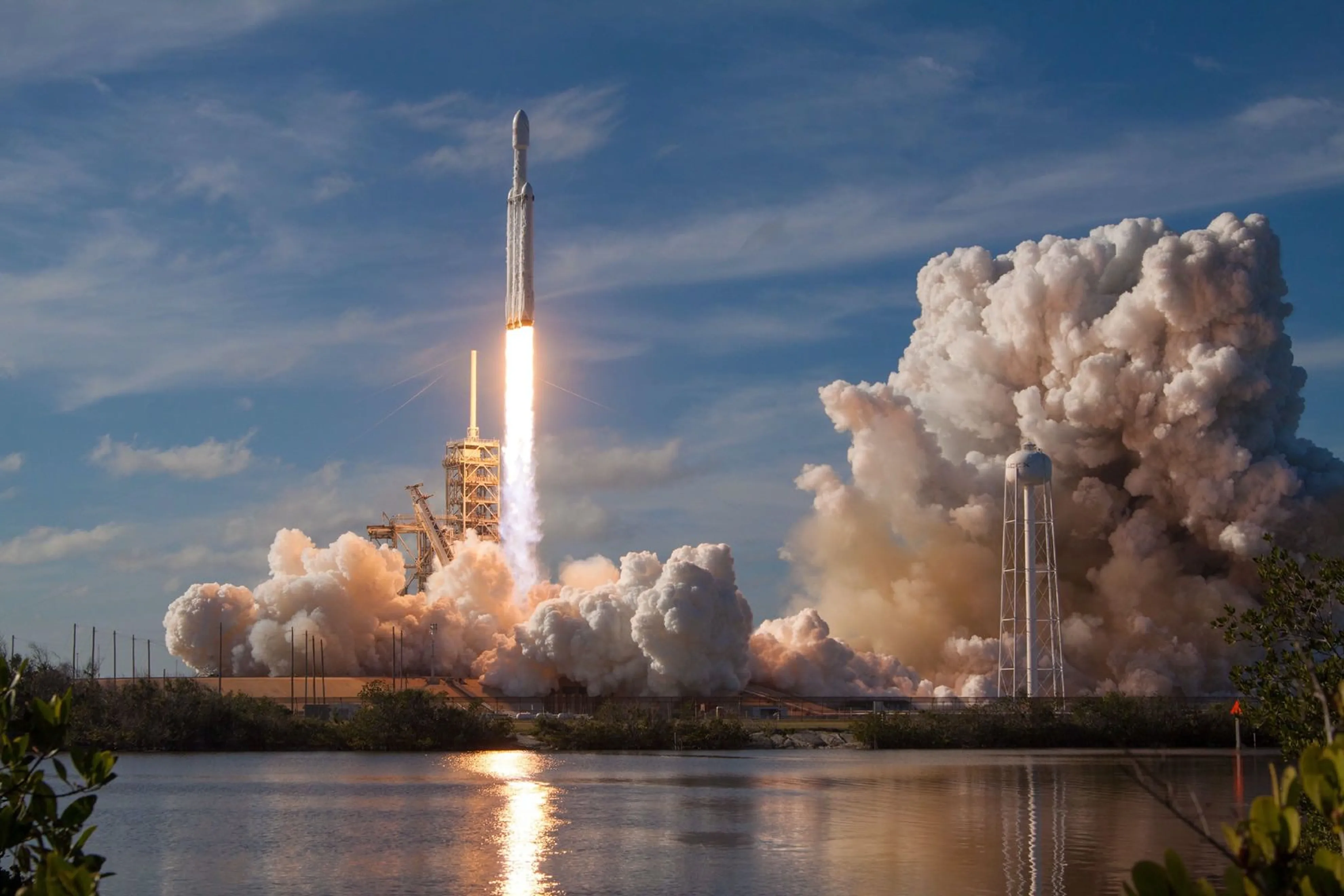 A rocket launching into the sky with bright flames and smoke clouds rising from the launch pad, reflected in the water below.